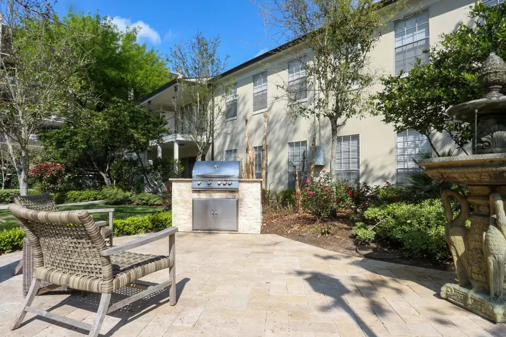 An outdoor sitting area with a fountain, 2 chairs, and a barbecue grill near Heritage Lofts apartment buildings, trees, and flowers.