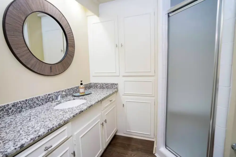 A bathroom with a stand up shower, a sink, large granite countertops, and ample cabinet and linen closet storage space.