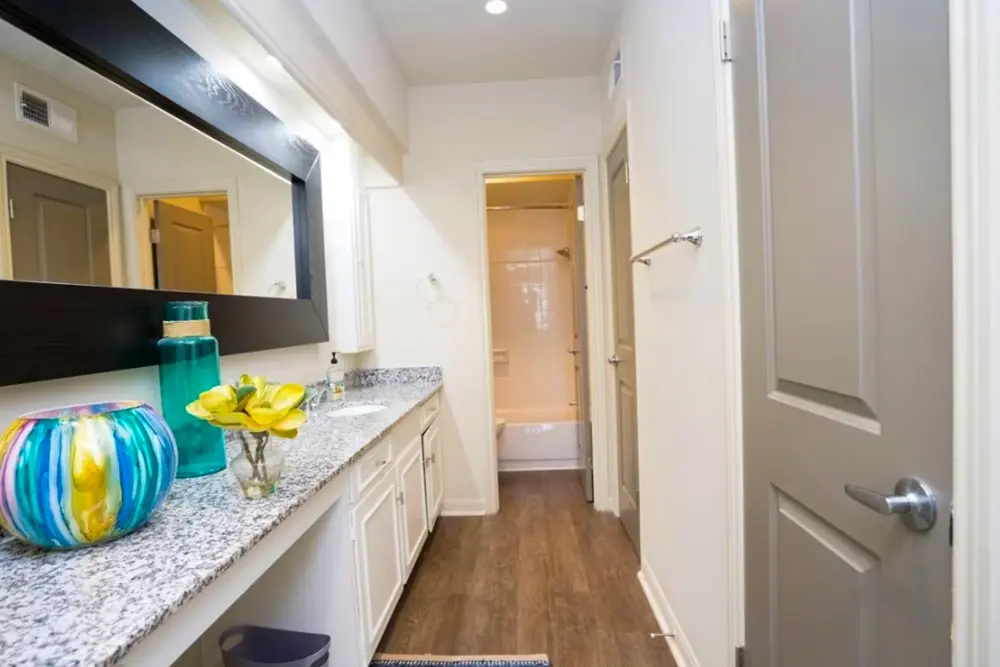 A long hallway with quartz countertops, a large mirror, under-the-sink cabinets leading to a bathroom with a shower and tub combination and a toilet.